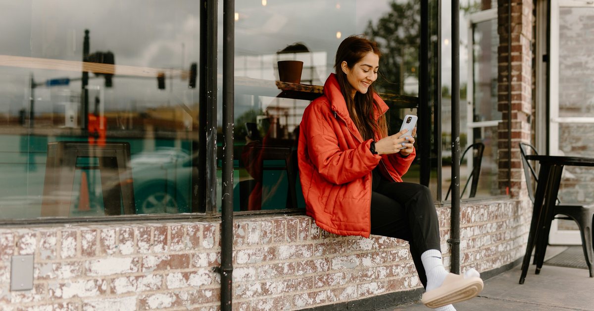 A woman in a red jacket on a café window ledge, smiling at her phone