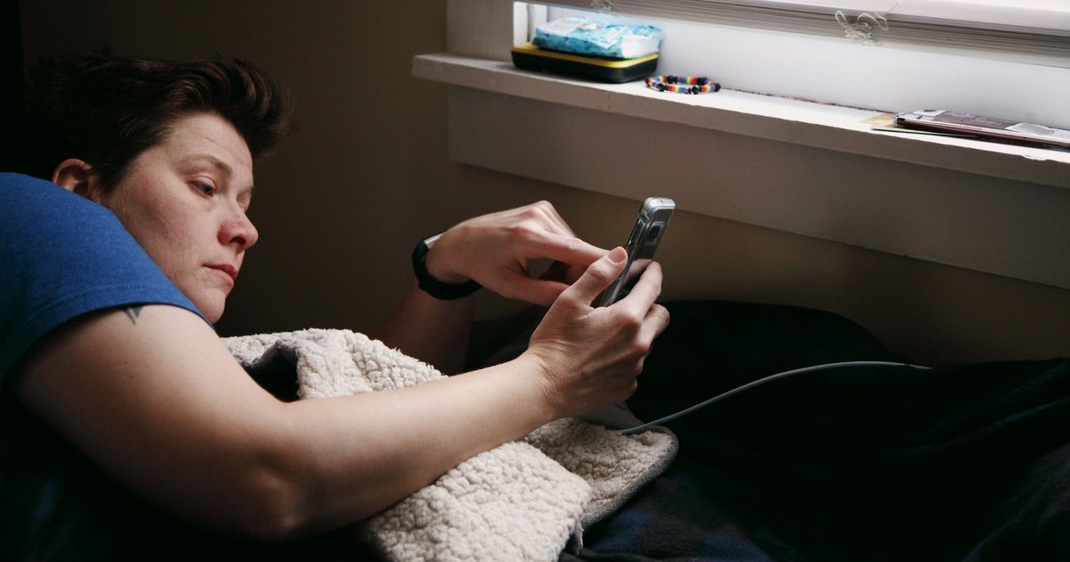 A man lying under a blanket, reading something on his phone