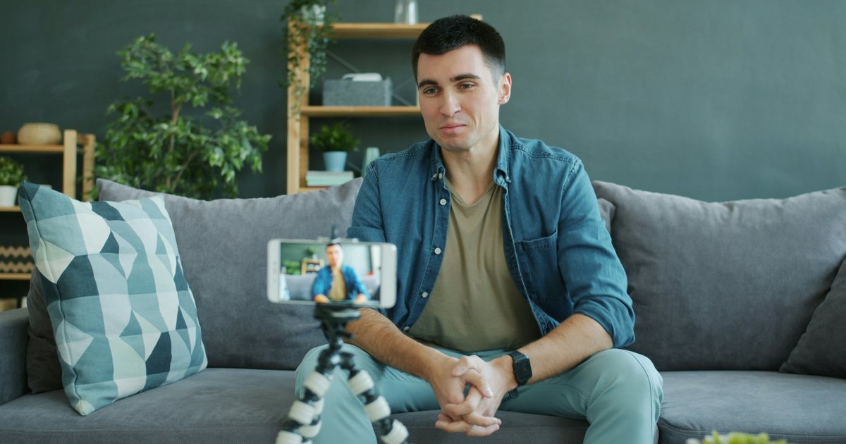 A man on a sofa recording himself with a phone on a small tripod